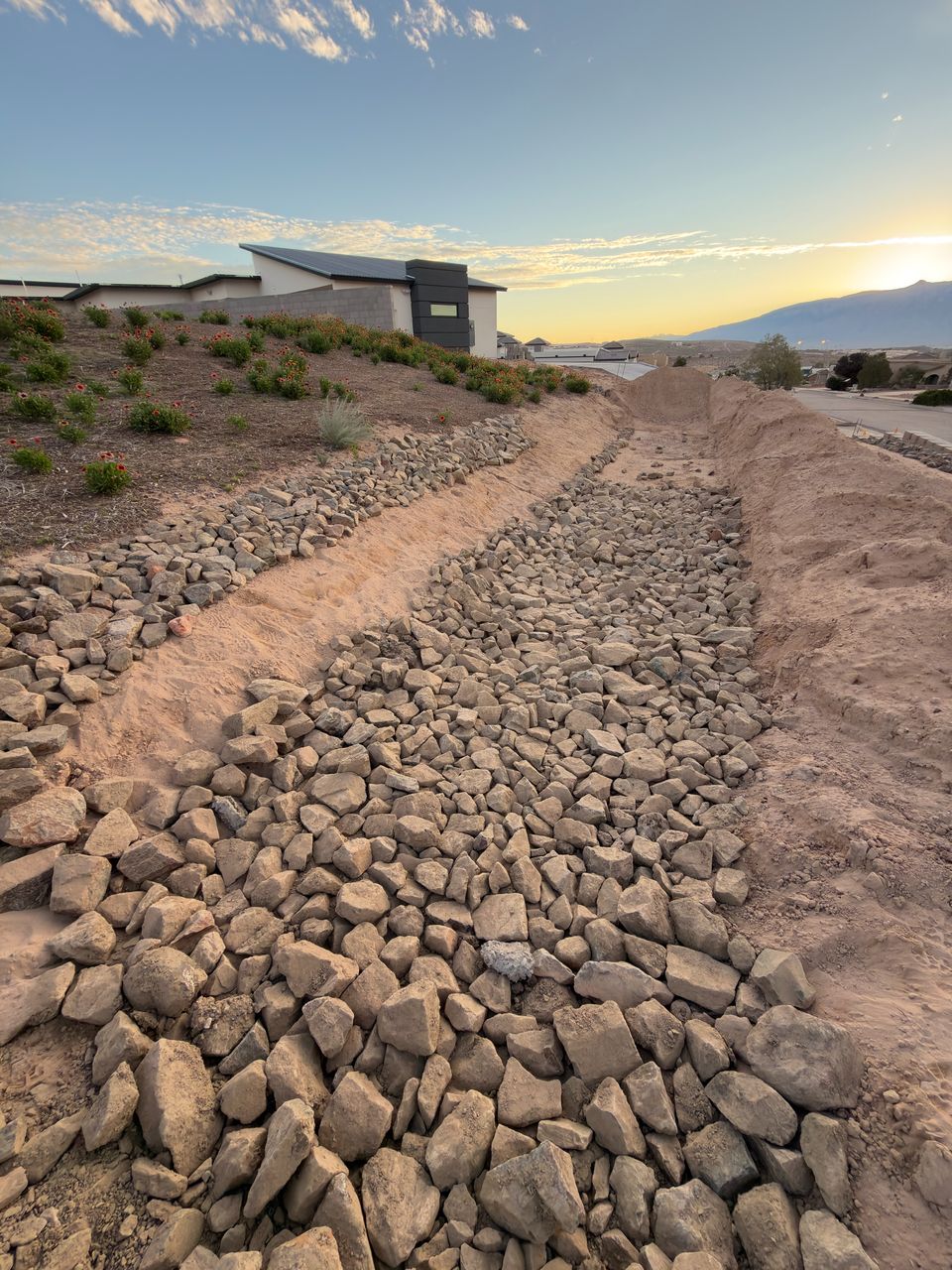 A finished rock-lined drainage channel running down a slope at sunset, the Sandia Mountains behind.