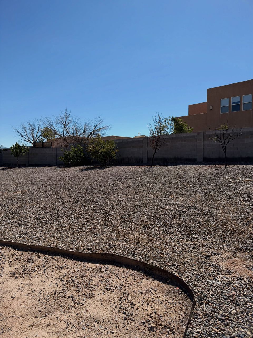 A large open gravel yard cleared of weeds, a block wall and houses behind.